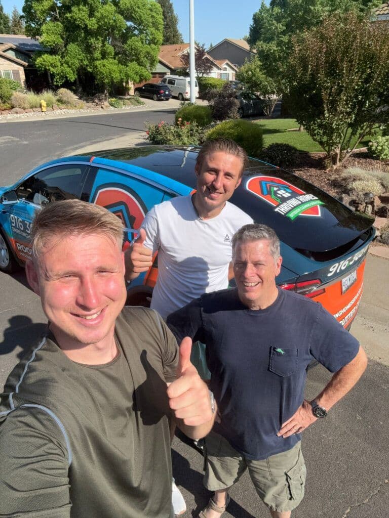 Three men giving thumbs up in front of a colorful branded car on a suburban street.